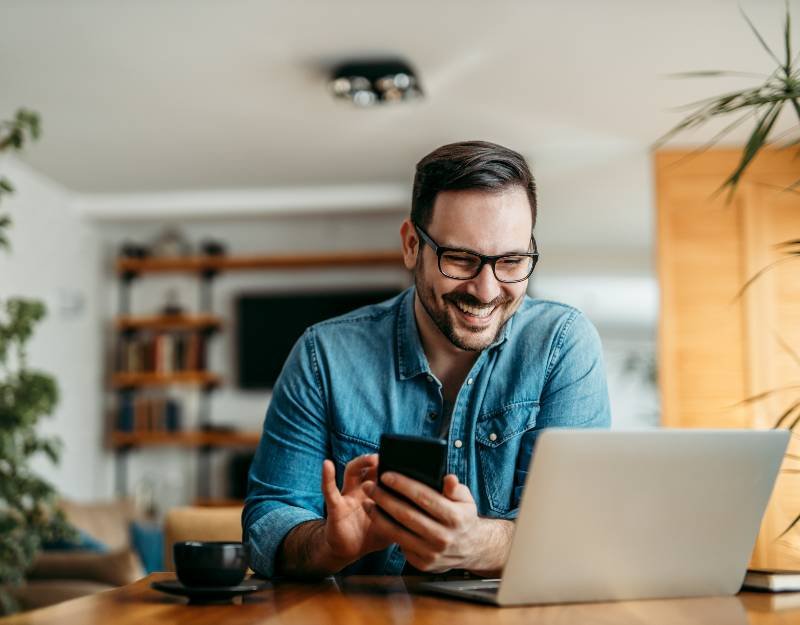 Smiling man in glasses using smartphone and laptop at home, illustrating digital service solutions and user engagement with WebHive software.